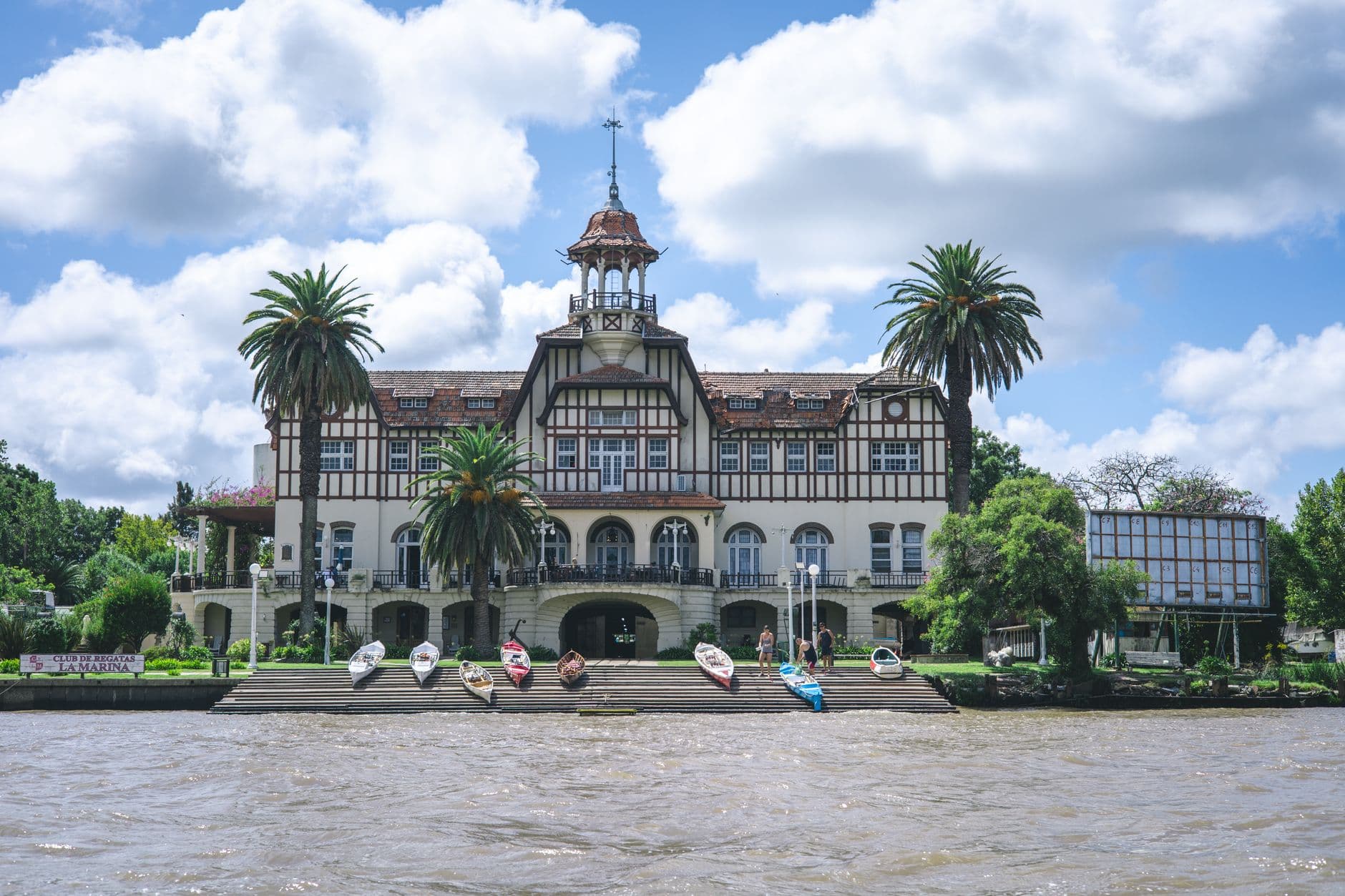 Tudor-style building with palm trees and boats by the water, under a bright blue sky.