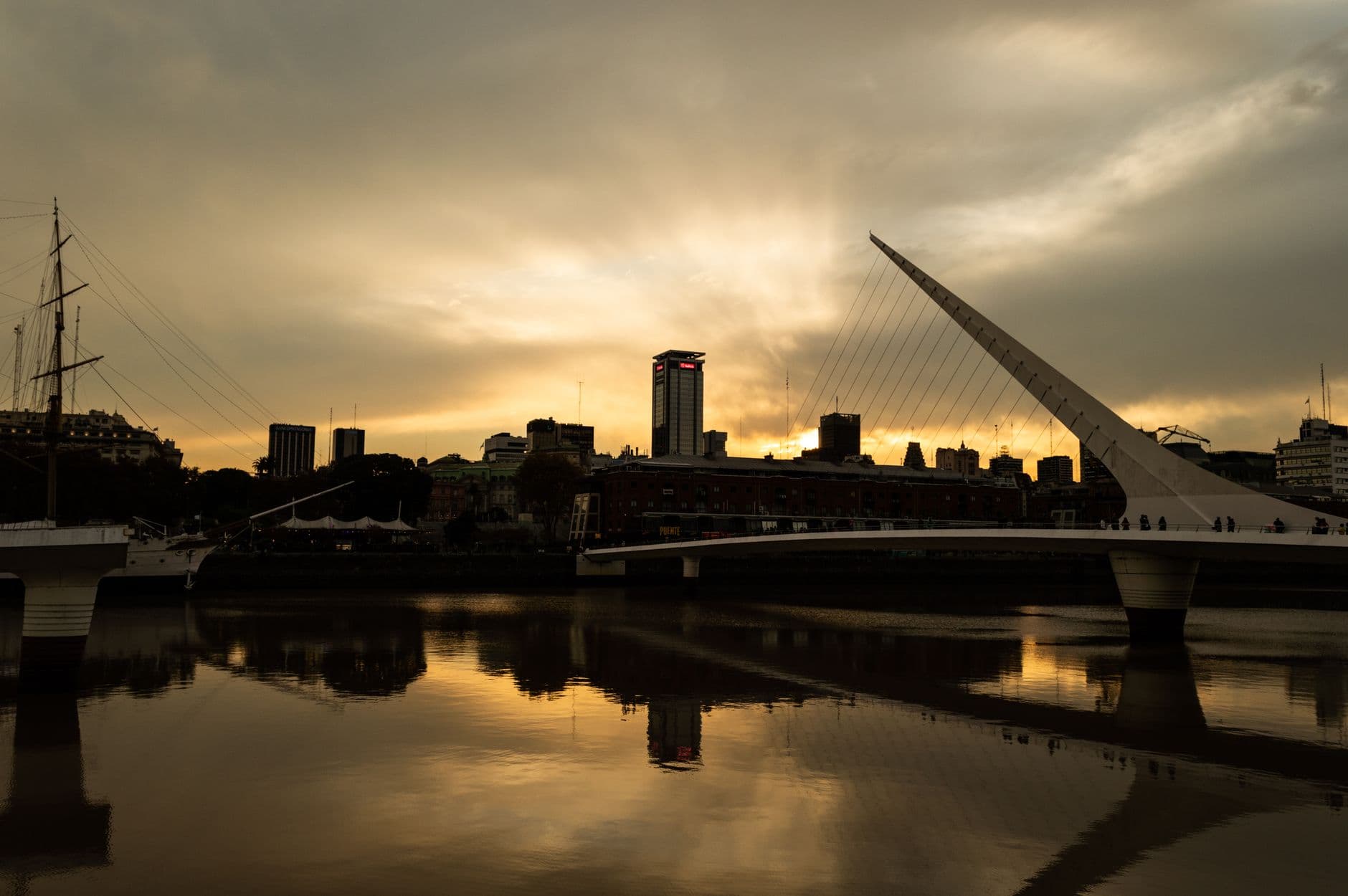 Silhouette of a city skyline with a modern bridge at sunset reflecting in calm waters.