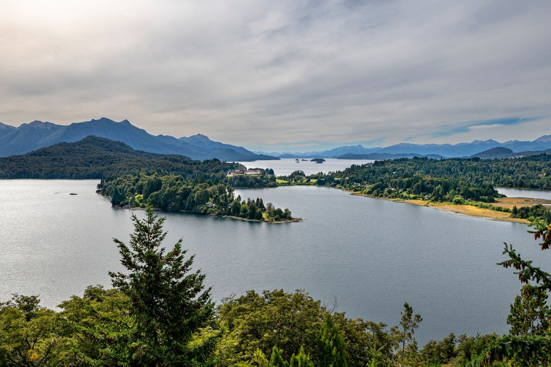 Scenic view of a serene lake surrounded by lush greenery and distant mountains under a cloudy sky.