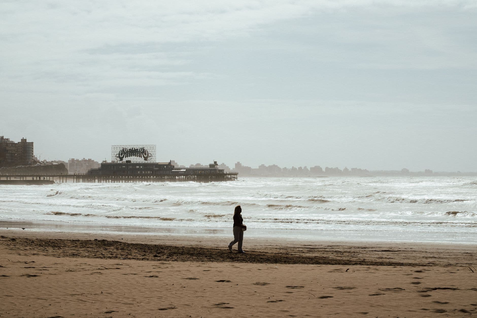 A lone person walks along a deserted beach with a vintage pier in the background.
