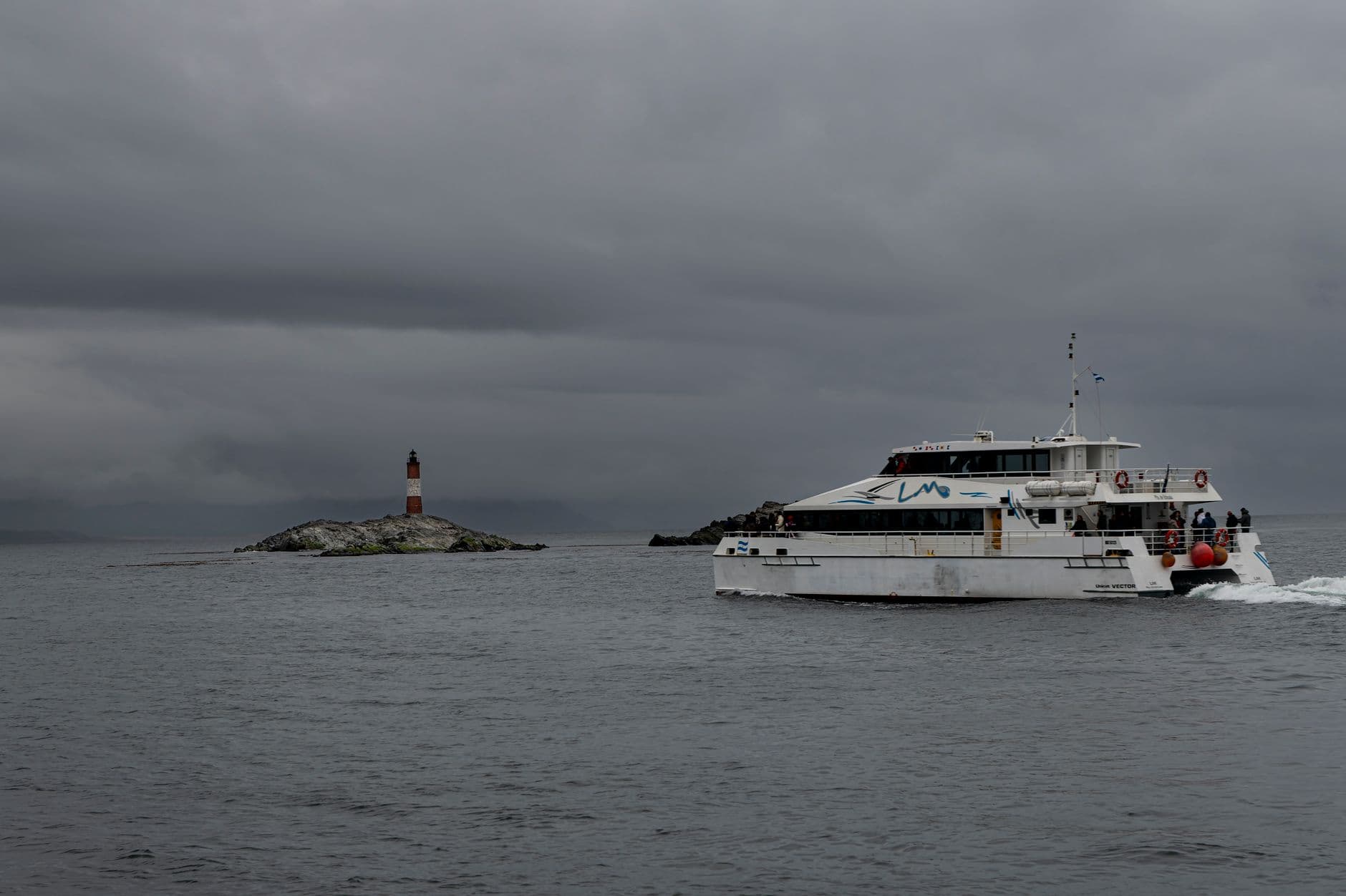 A catamaran sails near Les Eclaireurs Lighthouse under cloudy skies in Ushuaia, Argentina.
