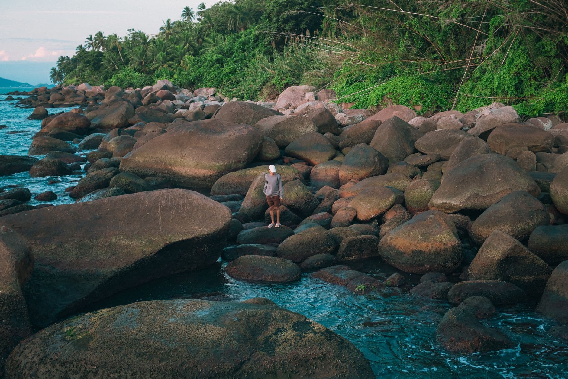 A lone figure stands on the rocky shore of Ilhabela, Brazil, surrounded by lush greenery.