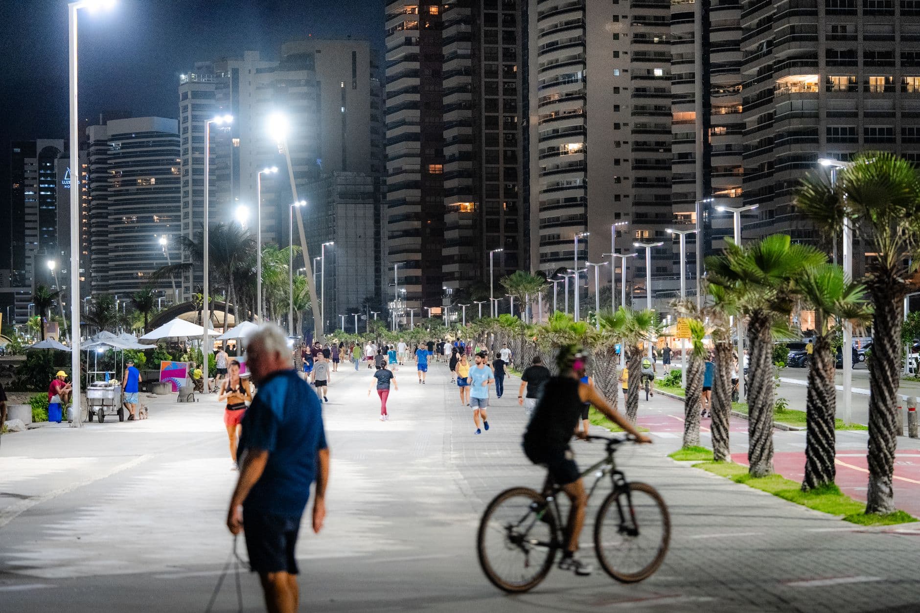 People enjoy night activities on a city promenade with illuminated buildings and palm trees.